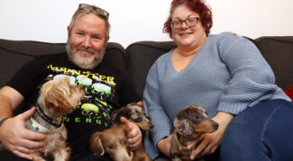 A man and woman sit together on a settee, both smiling and holding pet dogs