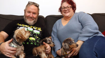 A man and woman sit together on a settee, both smiling and holding pet dogs