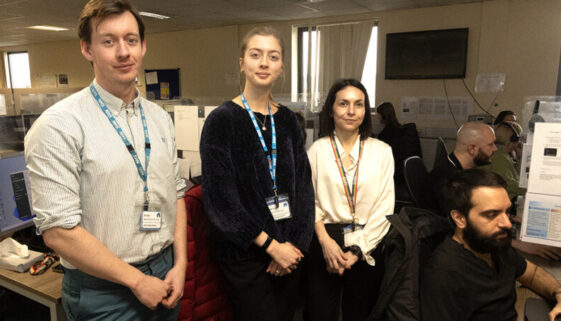 A man standing next two two women in an office call centre, with another man sitting at his desk