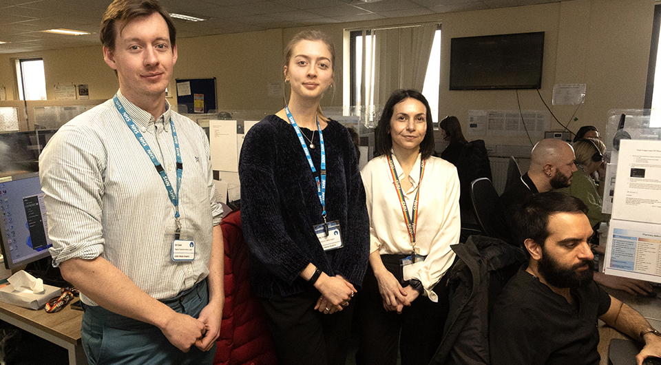 A man standing next two two women in an office call centre, with another man sitting at his desk